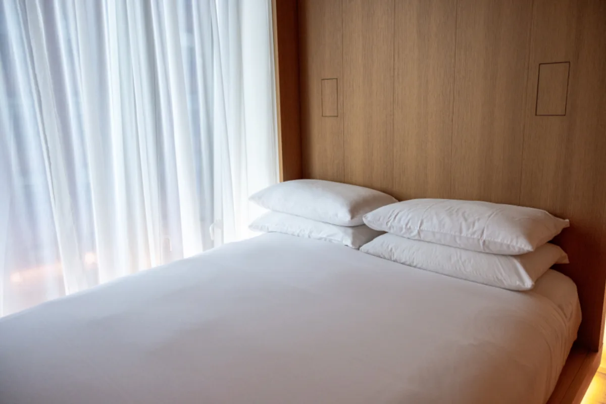 Bed made with white sheets and four white pillows against a wood panel wall, sheer white curtains beside it