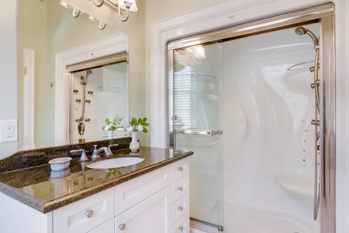Bathroom with a white vanity, a dark granite counter and a mirror, next to a sliding glass shower door in a silver frame with a white shower insert behind it