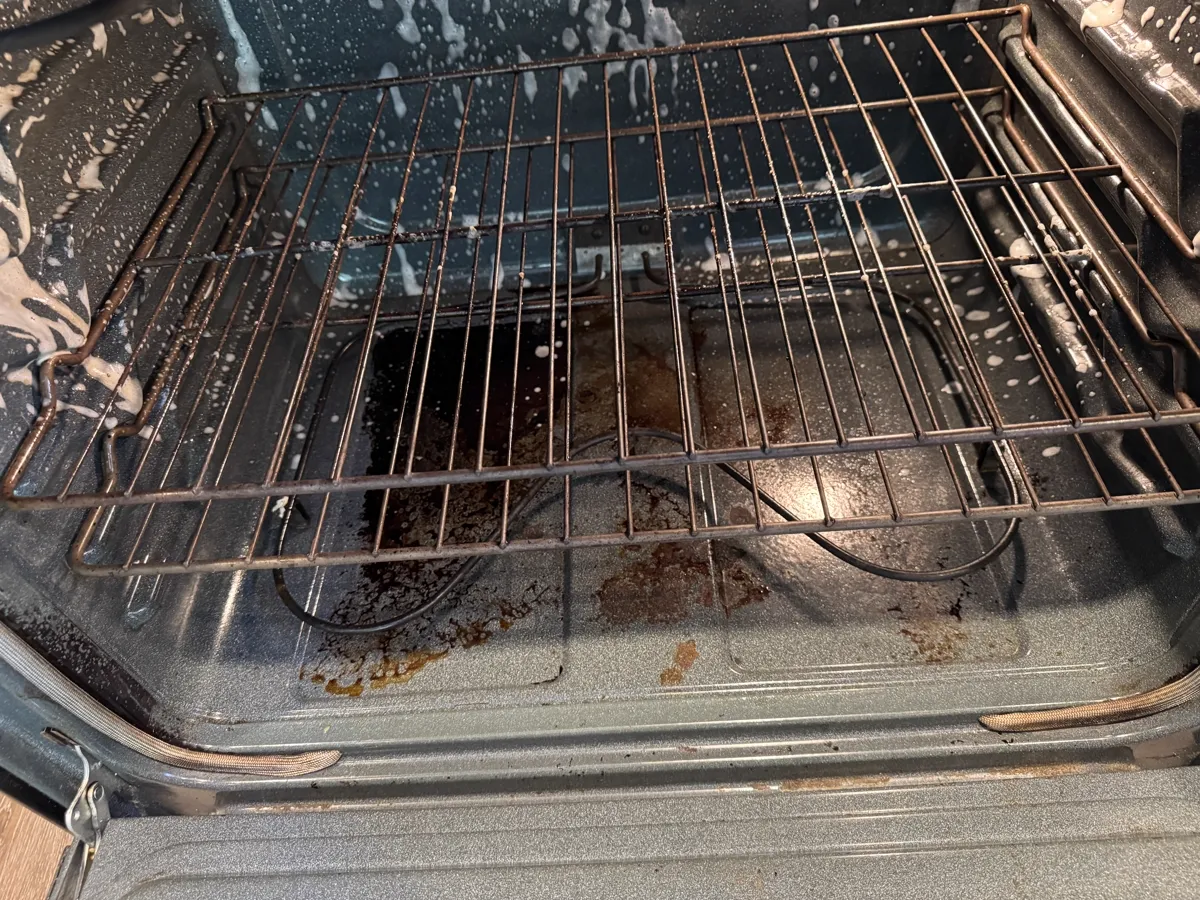 Oven interior with burnt food and grease on the floor, rack still in, cleaning foam on the walls