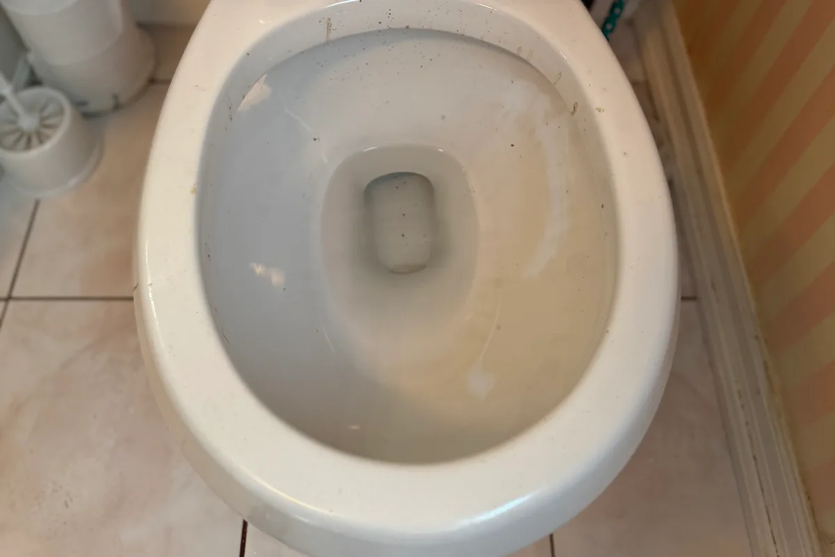 Toilet bowl and rim with dark spots and yellow stains, a white tiled floor around it