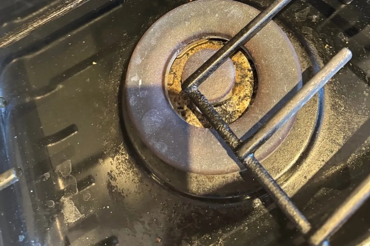 Close view of a gas burner cap and a black grate with crumbs, grease spots and residue around the burner