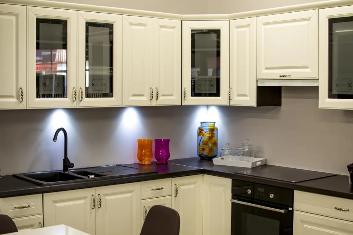 Cream kitchen cabinets with glass doors, lights under the upper cabinets, a grey backsplash and a dark counter with a black sink