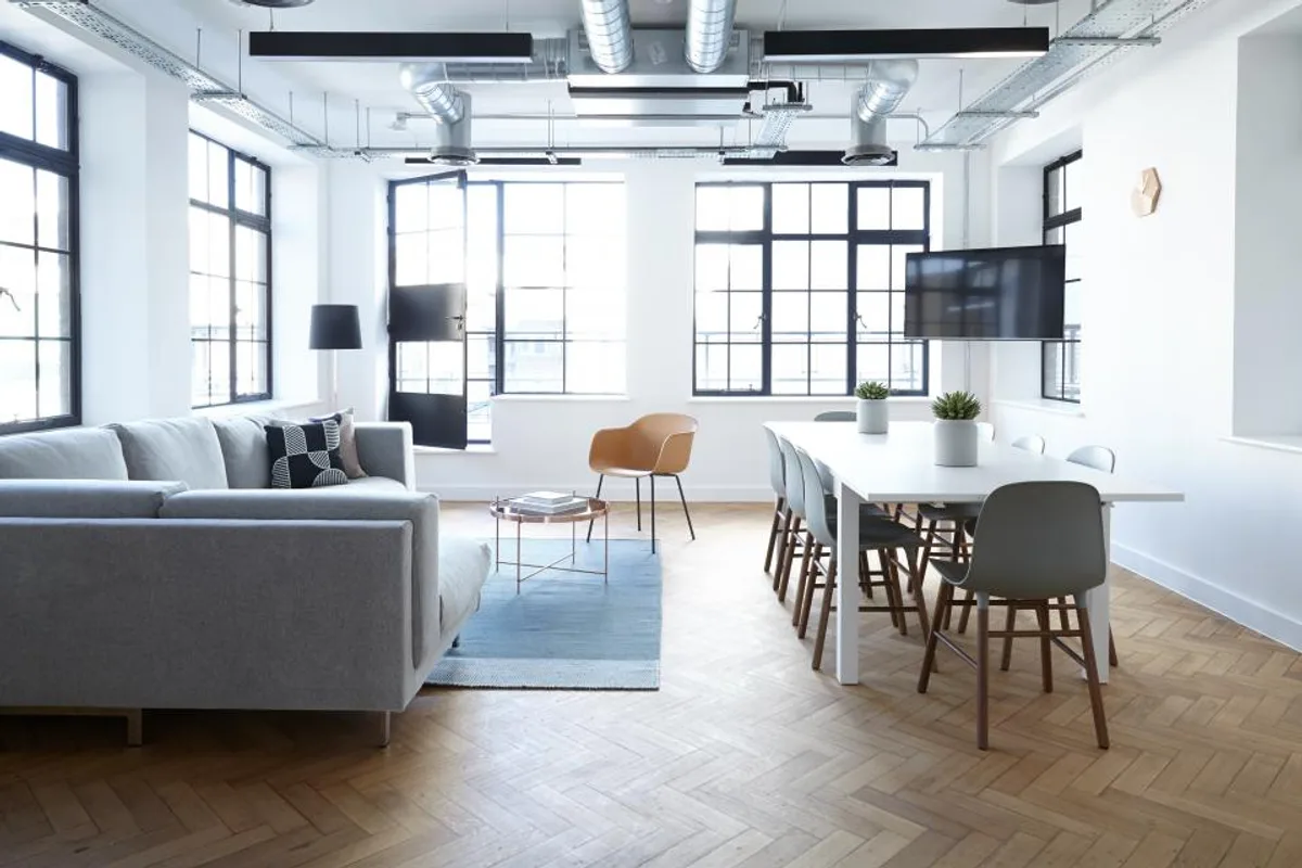 Bright open office with a grey sofa, a blue rug, a long white table with grey chairs and a herringbone wood floor