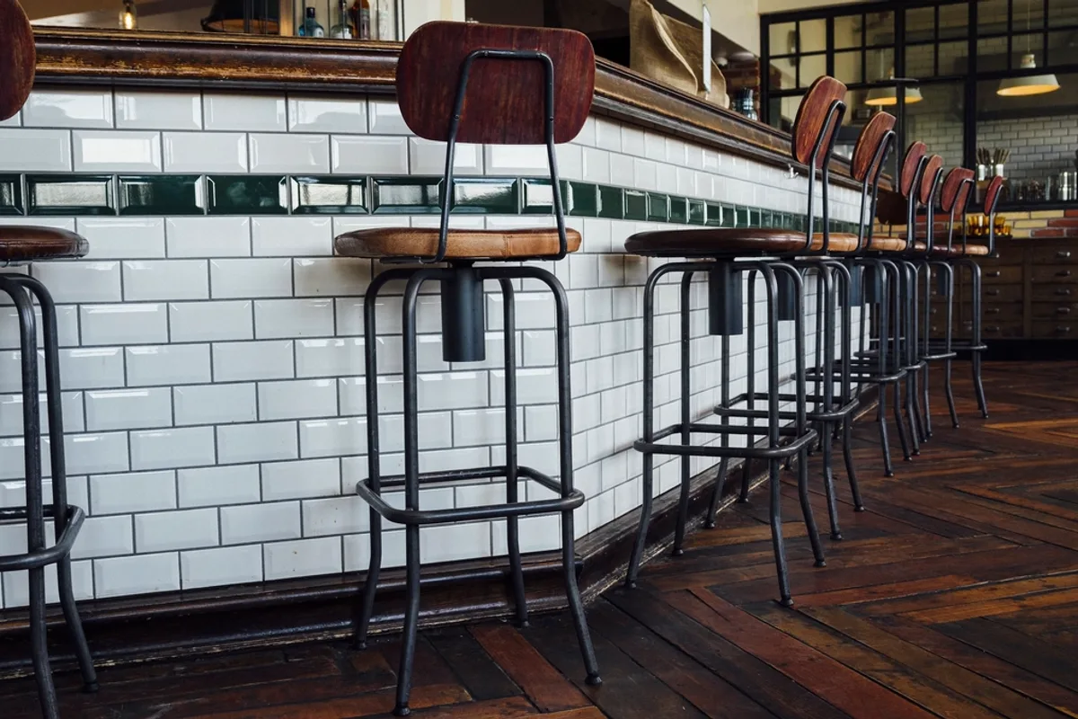 Row of bar stools with wooden seats and black metal legs against a white tiled bar on a dark herringbone wood floor