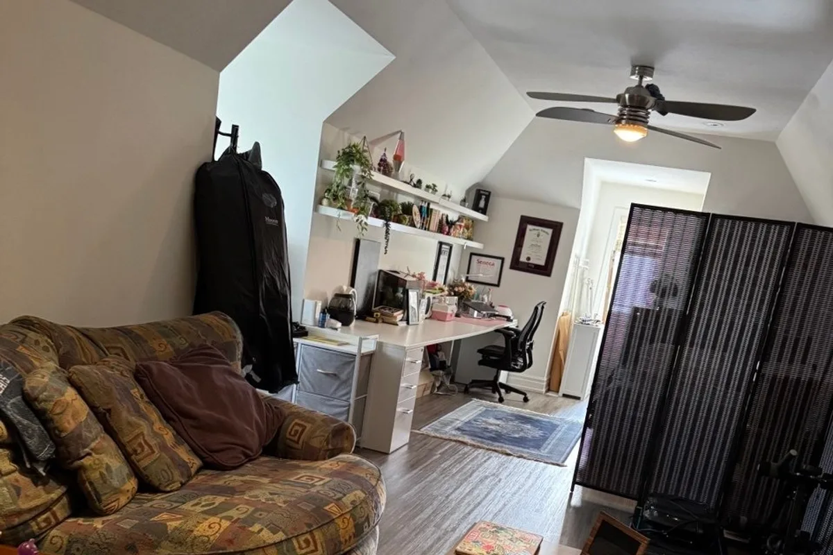 Upstairs room under a sloped ceiling with a patterned fabric sofa and cushions, a white desk, a room divider and a ceiling fan