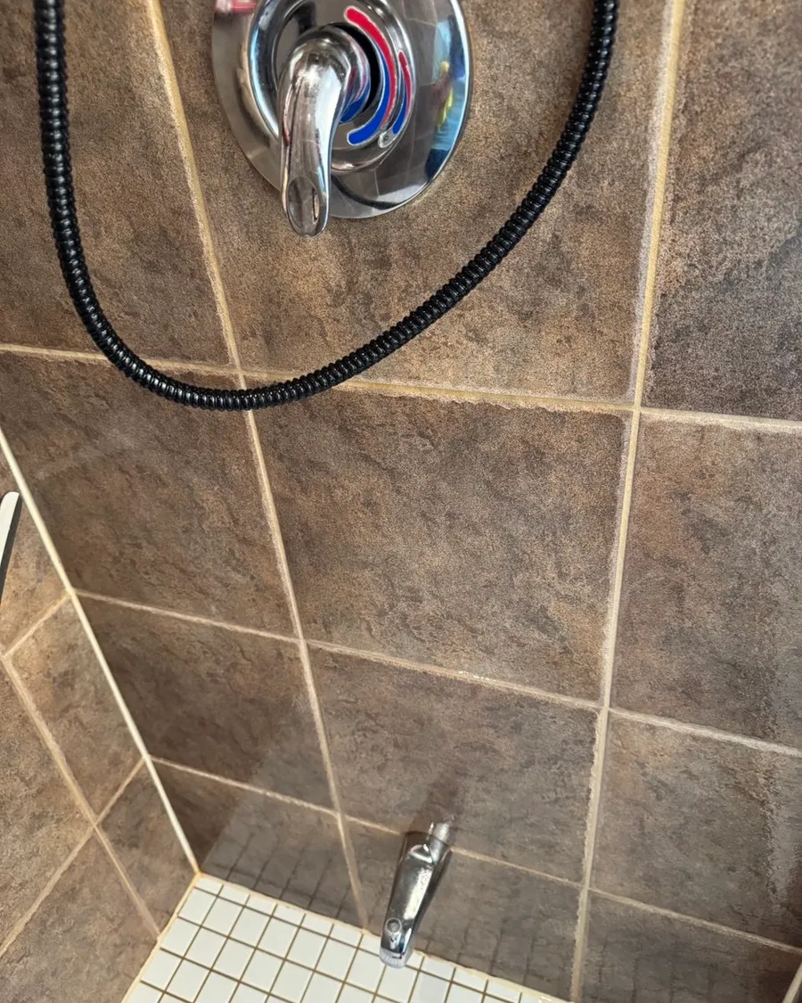 Shower wall of brown tile with a chrome handle, a spout and a white mosaic floor